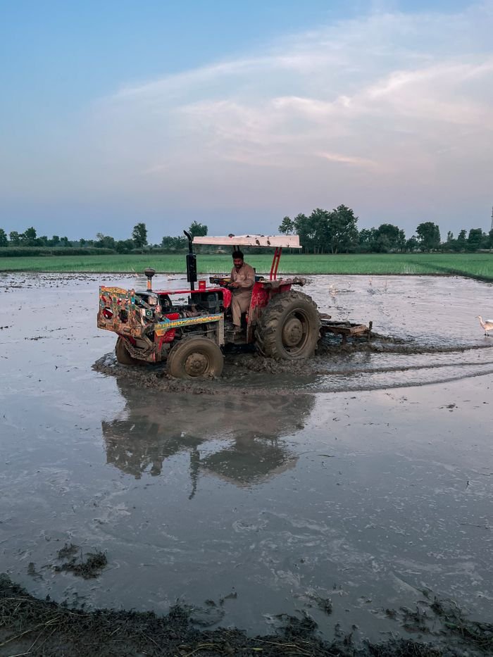 A tractor with a driver plowing a flooded field, reflecting in the water under a cloudy sky.